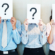 Three office workers lined up holding up pieces of paper in front of their faces with a question mark on them