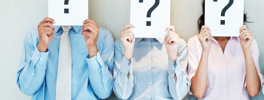 Three office workers lined up holding up pieces of paper in front of their faces with a question mark on them