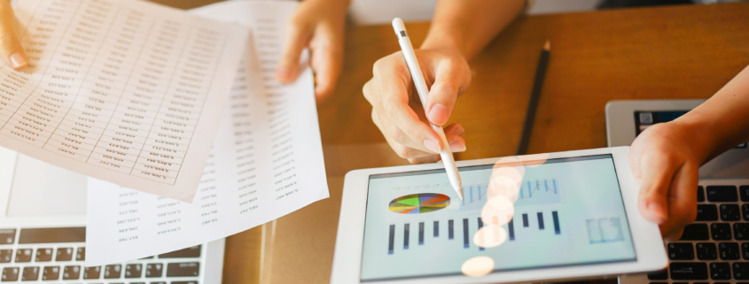 two employees working together, reviewing charts and numbers on a tablet and paperwork