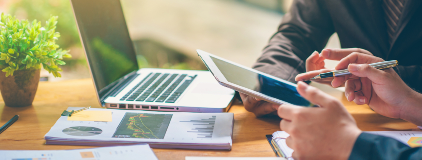 two employees converse over a tablet with documents and a laptop on a work desk